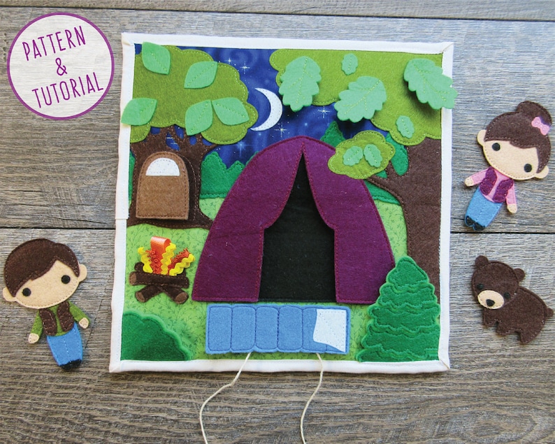 May include: A felt play set depicting a campsite with a purple tent, a campfire, a green tree, and two felt figures of people. The scene is set against a blue night sky with stars and a crescent moon.