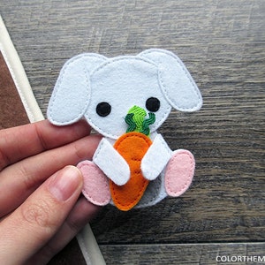 May include: A white felt bunny holding an orange carrot with green leafy tops. The bunny has black eyes and pink feet.