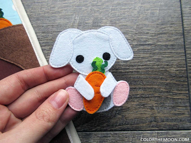 May include: A white felt bunny holding an orange carrot with green leafy tops. The bunny has black eyes and pink feet.