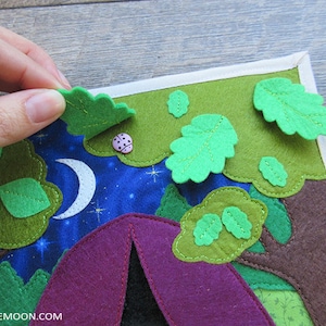 May include: A felt play mat with a purple tent, green trees, and a white crescent moon on a blue background. The mat has a white border and features a pink ladybug.