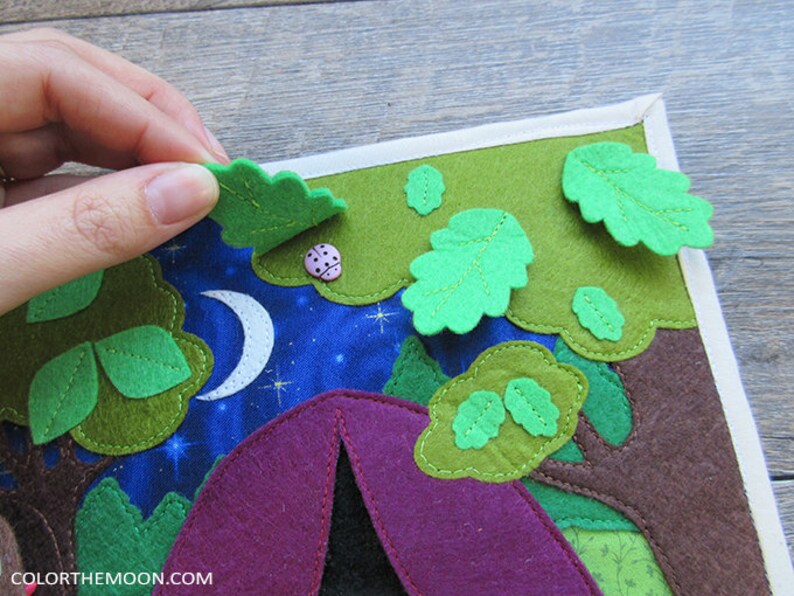 May include: A felt play mat with a purple tent, green trees, and a white crescent moon on a blue background. The mat has a white border and features a pink ladybug.
