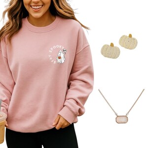 May include: A light pink sweatshirt with the text "Stay Spooky" and a ghost graphic. Two small, speckled, cream-colored pumpkin decorations and a gold necklace with a rectangular pendant are also shown.