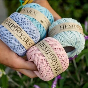 K&ouml;nnte beinhalten: Vier Kn&auml;uel Hanfgarn in Pastellfarben: Hellblau, T&uuml;rkis und Rosa. Jedes Kn&auml;uel hat ein braunes Papierband mit der Aufschrift "HEMP YARN". Das Garn wird in den H&auml;nden einer Person vor einem verschwommenen gr&uuml;nen und violetten Hintergrund gehalten.