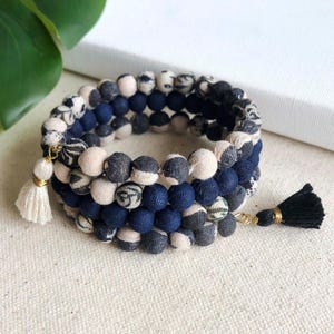 May include: A triple-strand bracelet made of fabric-covered beads in shades of blue and cream. The beads are patterned with a floral design and have gold accents. The bracelet has two tassels, one on each end.