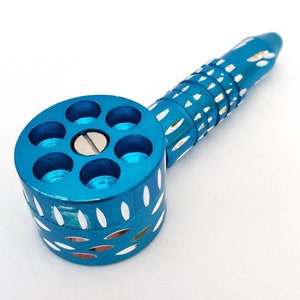 May include: A turquoise metal pipe with a revolver-style chamber and a cylindrical handle. The pipe features white decorative accents and is set against a white background. The chamber has six holes.