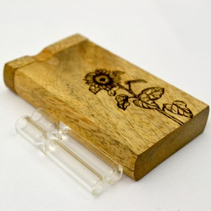 Può includere: Una scatola di legno con un disegno di girasole e due tubi di vetro. La scatola è in legno marrone chiaro e ha una finitura naturale. Il disegno del girasole è bruciato nel legno ed è di colore marrone scuro. I tubi di vetro sono trasparenti e hanno una finitura liscia.