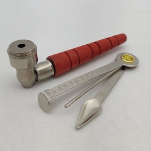 May include: A red and silver smoking pipe with a metal tamper and a small metal poker. The tamper has a decorative pattern.