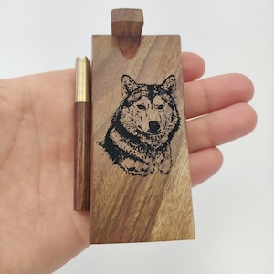 May include: A wooden box with a wolf design engraved on the front. The box has a small wooden lid and a brass-colored metal insert. The box is in the hand of a person.