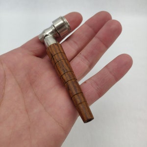 Könnte beinhalten: Eine kleine Pfeife mit einem Holzkörper und einer Metallschale. Der Holzteil ist dunkelbraun mit einem gestreiften Muster. Die Metallschale ist silberfarben und abgewinkelt.