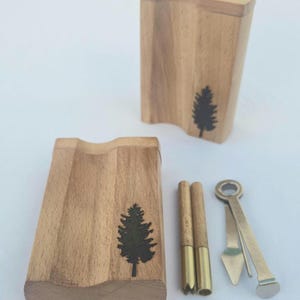 May include: Wooden box set with a carved design and a black tree silhouette. Includes two wooden tubes with brass tips and a metal tool. The set is designed for a specific purpose.