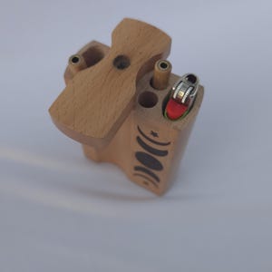 May include: A wooden box with a hinged lid, featuring a lighter and several cylindrical holes. The box is light brown with a decorative black crescent moon and star design on the side. The lighter has a red top.