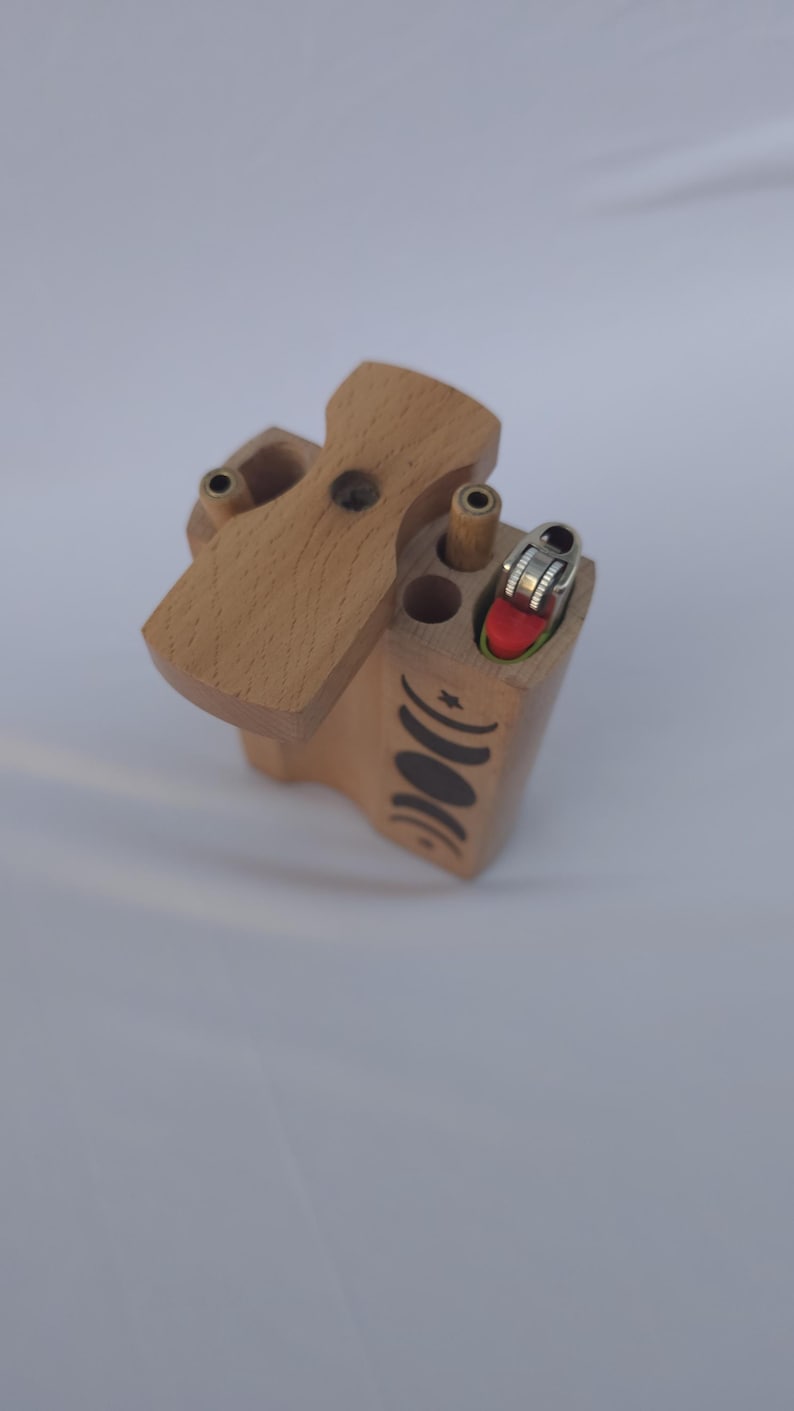 May include: A wooden box with a hinged lid, featuring a lighter and several cylindrical holes. The box is light brown with a decorative black crescent moon and star design on the side. The lighter has a red top.