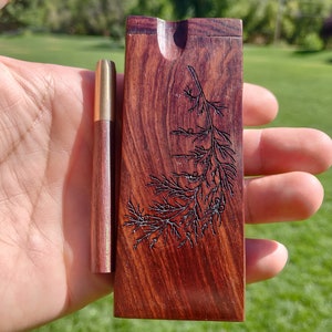May include: A wooden box with a carved design of a leafy branch. The box has a small opening on the top and is made of dark red wood. A small wooden pipe is next to the box.