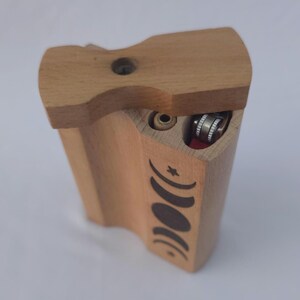 May include: A wooden dugout box with a sliding lid. The box is light brown with a carved design of moon phases. It has a compartment for a lighter and a small hole. The lighter is silver and red.