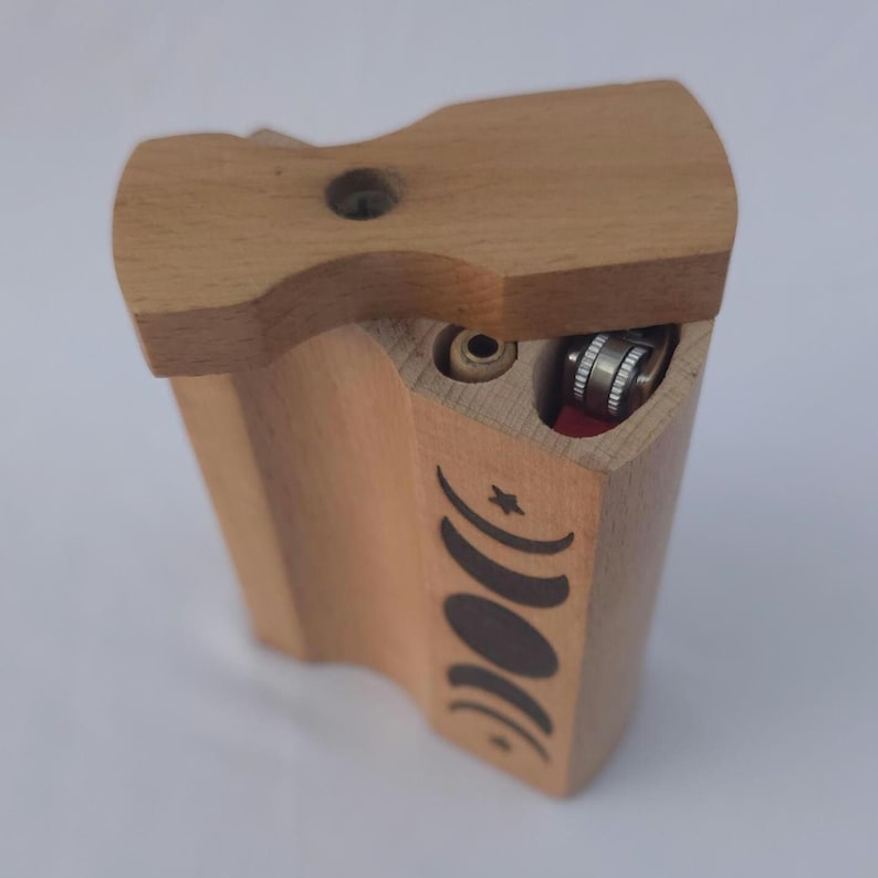May include: A wooden dugout box with a sliding lid. The box is light brown with a carved design of moon phases. It has a compartment for a lighter and a small hole. The lighter is silver and red.