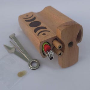 May include: A wooden box with a carved moon phase design, holding a lighter and small wooden tubes. A metal tool and a small bag are next to the box. The box is made of light-colored wood.