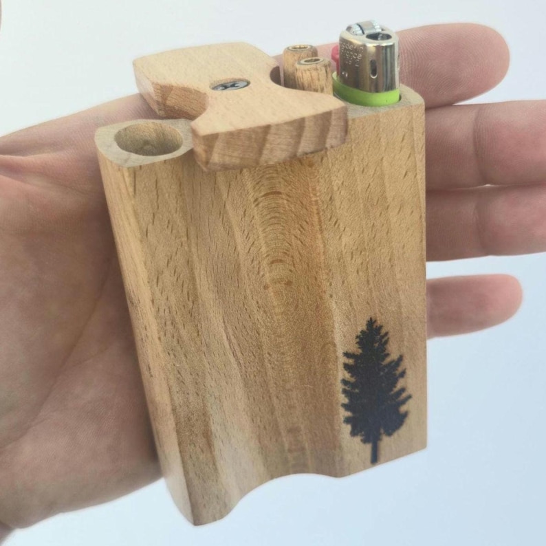 May include: A wooden dugout with a lighter and a small compartment. The dugout is made of light-colored wood and features a black tree graphic on the side. The lighter is green and silver.