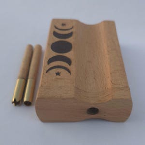May include: A light-colored wooden box with a carved groove and a series of moon phase designs. Two wooden tools with brass tips are next to the box. The box has a small hole on the side.