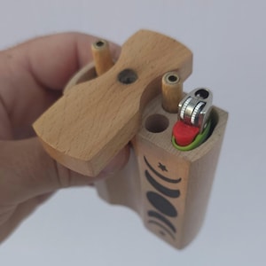 May include: A light-colored wooden dugout with a hinged lid. The dugout has a compartment for a lighter and a decorative black moon phase design. A lighter with a red and green base is visible.