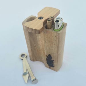 May include: A wooden box with a hinged lid, featuring a small tree graphic. It contains a lighter and two cylindrical openings. A metal tool rests nearby. The box is made of light-colored wood.