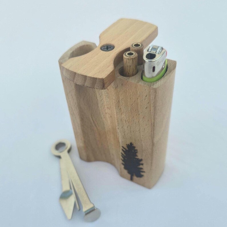 May include: A wooden box with a hinged lid, featuring a small tree graphic. It contains a lighter and two cylindrical openings. A metal tool rests nearby. The box is made of light-colored wood.