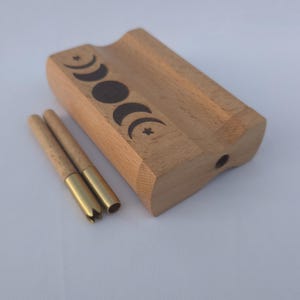 May include: A wooden box with a carved groove and a series of moon phase designs. Two cylindrical tools with brass tips are next to the box. The box and tools are made of wood.