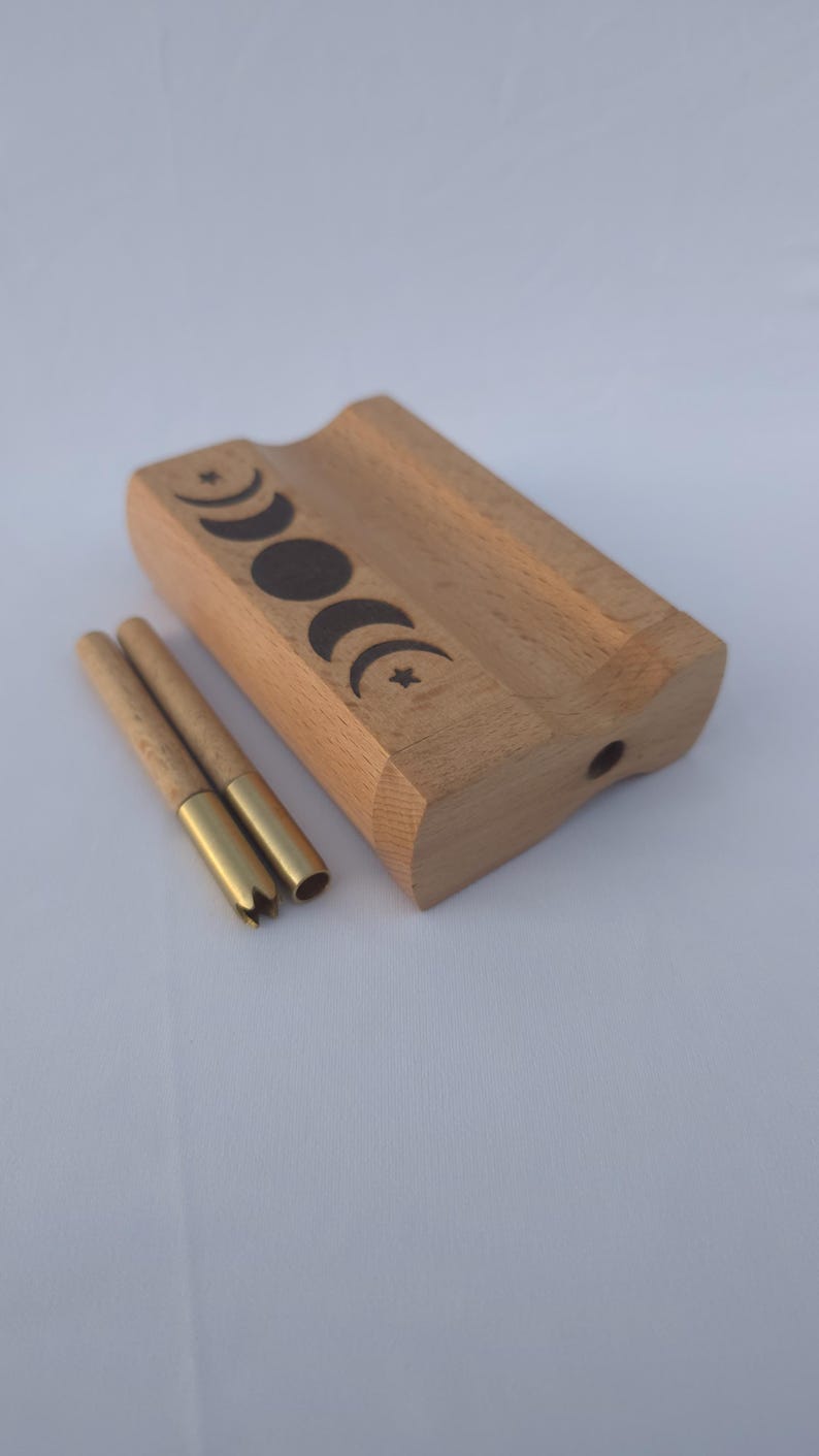 May include: A wooden box with a carved groove and a series of moon phase designs. Two cylindrical tools with brass tips are next to the box. The box and tools are made of wood.