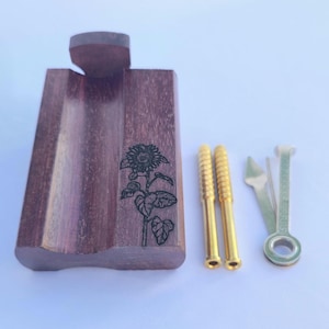 May include: A wooden pipe with a sunflower engraving, accompanied by two gold-colored metal pipe screens and a pipe tool. The pipe is a dark brown color, and the sunflower is etched into the wood.