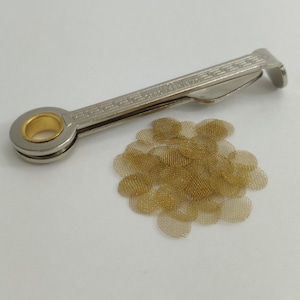 Puede incluir: Una herramienta de pipa de plata con un tampón incorporado y un agujero redondo para sujetar una pantalla. Un montón de pantallas de pipa de metal dorado se encuentran en el fondo.