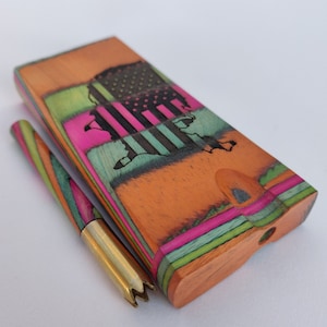 May include: A wooden box and matching tool with a colorful layered design. The box features an etched American flag design with pink, green, and black accents. The tool has a gold-colored tip.