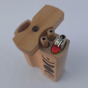 May include: A wooden dugout box with a hinged lid, featuring a lighter and two cylindrical compartments. The box is light brown with a black crescent moon design on the side. The lighter has a red button.