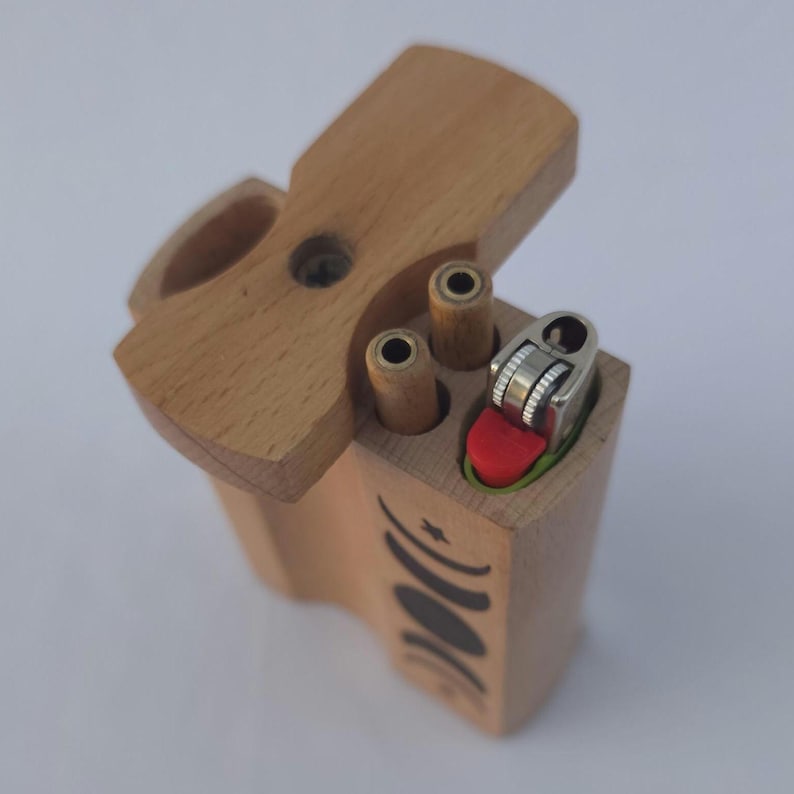 May include: A wooden dugout box with a hinged lid, featuring a lighter and two cylindrical compartments. The box is light brown with a black crescent moon design on the side. The lighter has a red button.