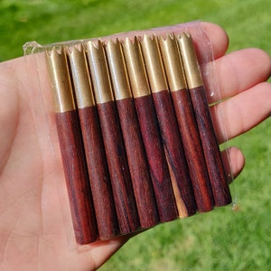 May include: A set of ten wooden dowels with gold metal tips. The dowels are a dark reddish brown color and have a wood grain pattern. The gold tips are pointed and have a decorative design.
