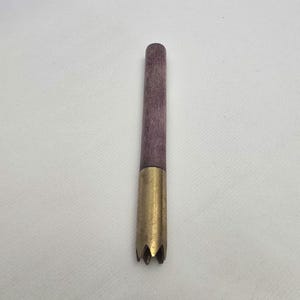 Puede incluir: Una herramienta de madera púrpura con una punta de latón. La punta de latón tiene un diseño en forma de estrella. La herramienta está diseñada para una artesanía o pasatiempo específico. El mango de madera es de color púrpura apagado.