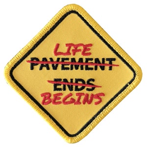 Puede incluir: Parche amarillo en forma de diamante con borde negro y costuras rojas. El texto "Life Begins" está escrito en rojo con la palabra "Pavement" tachada en rojo. El texto "Ends" también está tachado en rojo.