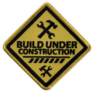 Puede incluir: Parche amarillo en forma de diamante con borde negro y texto negro que dice "BUILD UNDER CONSTRUCTION". El parche tiene un símbolo negro de llave inglesa y martillo en la parte superior e inferior.