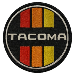 May include: A black and white embroidered patch with the word "TACOMA" in white lettering and a design of four vertical stripes in red, orange, and yellow.