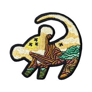 Puede incluir: Parche bordado con forma de cachorro de león, con un diseño de paisaje. El parche tiene un borde negro y muestra una escena de puesta de sol con árboles, montañas y un cielo amarillo. El diseño está detallado con varios colores.