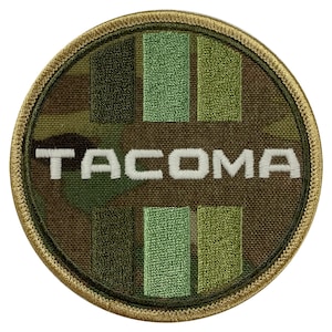 Könnte beinhalten: Ein runder, brauner Patch mit einem Tarnmuster. Der Patch hat das Wort "TACOMA" in weiß gestickt in der Mitte. Das Tarnmuster besteht aus grünen, braunen und beigen Farben.