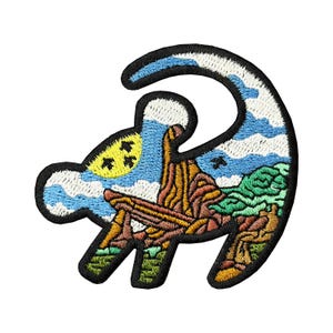 May include: Embroidered patch in the shape of a lion cub's head, featuring a landscape scene. The design includes a blue sky with clouds, a yellow sun, and a mountain range with green foliage. The patch is outlined in black.