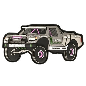 Puede incluir: Un camión de juguete gris y negro con detalles en rosa. El camión tiene los logotipos "TOYOTIRES" y "MONSTER ENERGY" en el lateral. El camión es una réplica de una Toyota Tundra.