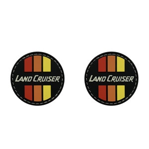 Könnte beinhalten: Zwei runde schwarze Patches mit weißen Nähten an den Rändern. Jeder Patch hat ein rot-orange-gelbes Streifenmuster mit dem Text "LAND CRUISER" in Weiß.
