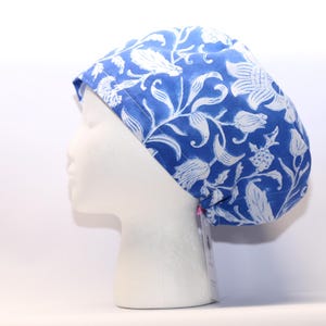 May include: A blue and white floral patterned scrub cap. The cap features a repeating design of stylized flowers and leaves. The fabric is a vibrant blue with the floral pattern in white. The cap is shown on a white mannequin head.