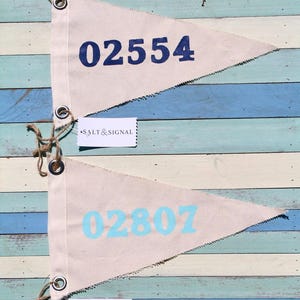 May include: Two triangular canvas pennants with metal grommets and jute twine. The top pennant features the dark blue number 02554, and the bottom pennant features the light blue number 02807. Each pennant includes a small white tag labeled SALT & SIGNAL.