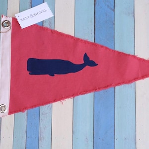 May include: A triangular red pennant flag featuring a dark blue silhouette of a whale in the center. The flag has a white vertical strip on the left side with two metal grommets and attached twine. A white tag reads SALT & SIGNAL.