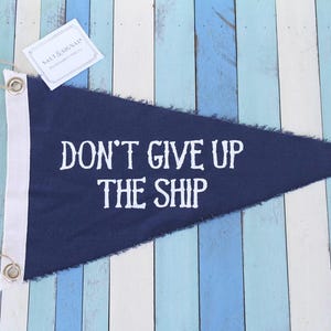 以下が含まれることがあります： DON'T GIVE UP THE SHIPという白い文字がプリントされた、紺色の三角形の布製ペナント旗。旗には2つの金属製ハトメと紐が付いた白いキャンバス地のヘッダーがあり、SALT & SIGNALと書かれた小さな白い長方形のタグが付いています。