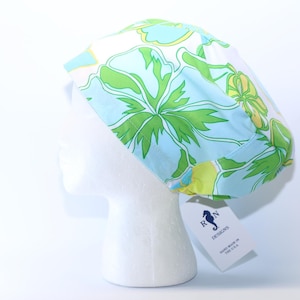 May include: A floral patterned scrub cap in shades of green, blue, and yellow, displayed on a white mannequin head. The cap features a vibrant botanical design. A tag attached to the cap reads "R N DESIGNS HAND MADE IN THE U.S.A."