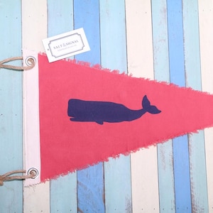 Op de afbeelding: Een driehoekige rode wimpel met een donkerblauw silhouet van een walvis in het midden. De vlag heeft een witte verticale strook aan de linkerkant met twee metalen ringen en bevestigd jutekoord. Een wit label met de tekst SALT & SIGNAL is bevestigd.
