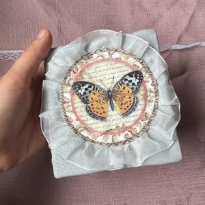 May include: A square light gray fabric box featuring a circular decorative topper. The topper displays a butterfly illustration with orange and black wings, surrounded by a ruffled white sheer ribbon trim and a vintage-style script background.