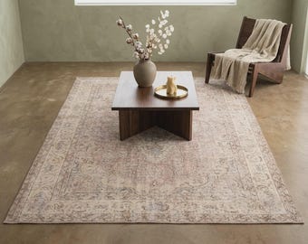 May include: A rectangular area rug featuring a faded, intricate floral pattern in muted shades of beige, dusty rose, and slate blue. The rug is placed under a dark wood square coffee table holding a ceramic vase with cotton stems and a small gold tray.
